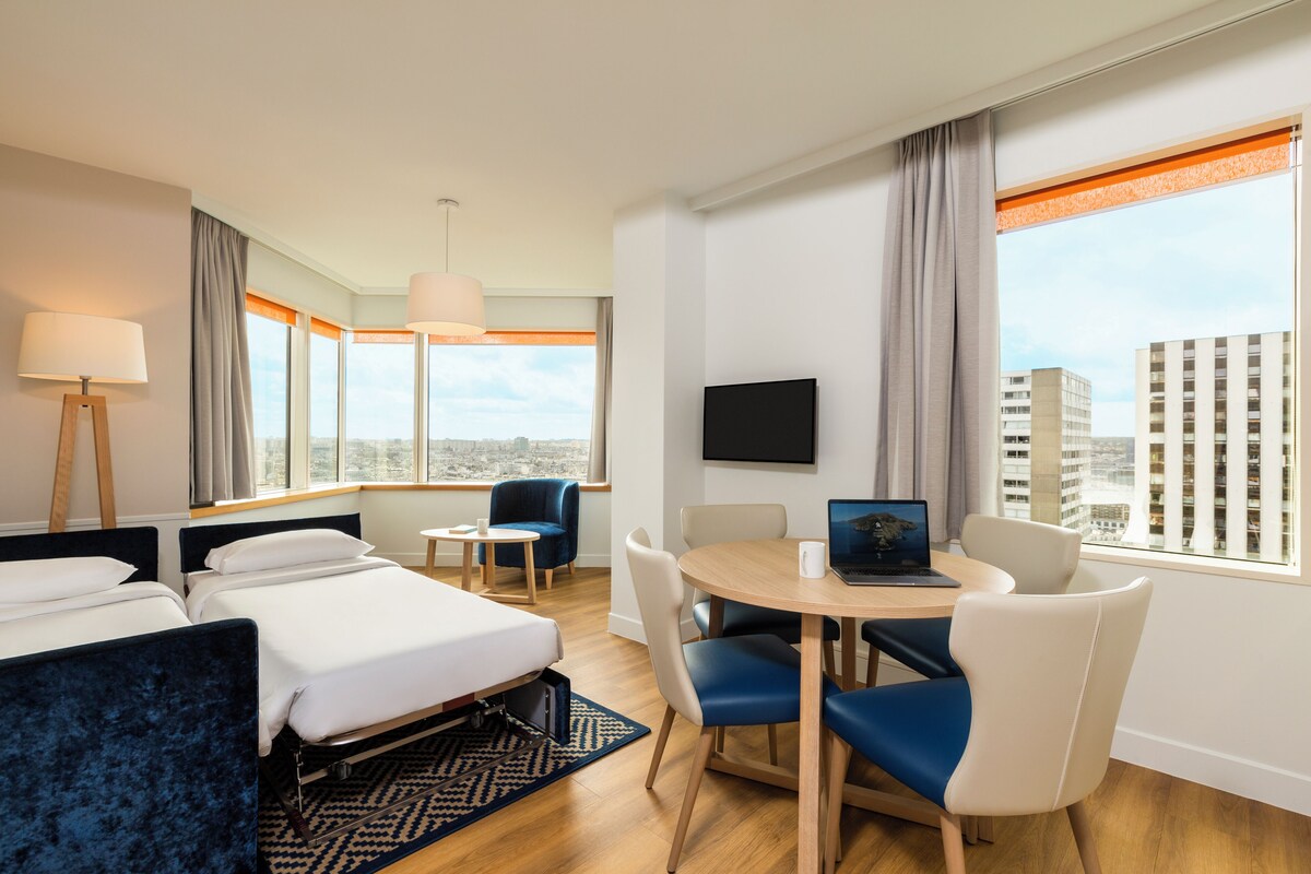Habitación de hotel con dos camas, un escritorio con una computadora portátil, televisión y vista a la ciudad.