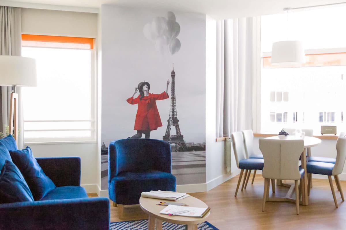 Habitación de hotel con un gran mural de la Torre Eiffel, un sofá azul, una mesa de comedor con sillas y una mesita de café.