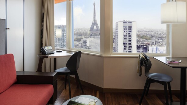 Una habitación de hotel con vistas a la Torre Eiffel, un escritorio con una lámpara, una silla, un sofá rojo y una mesita con un vaso de jugo.