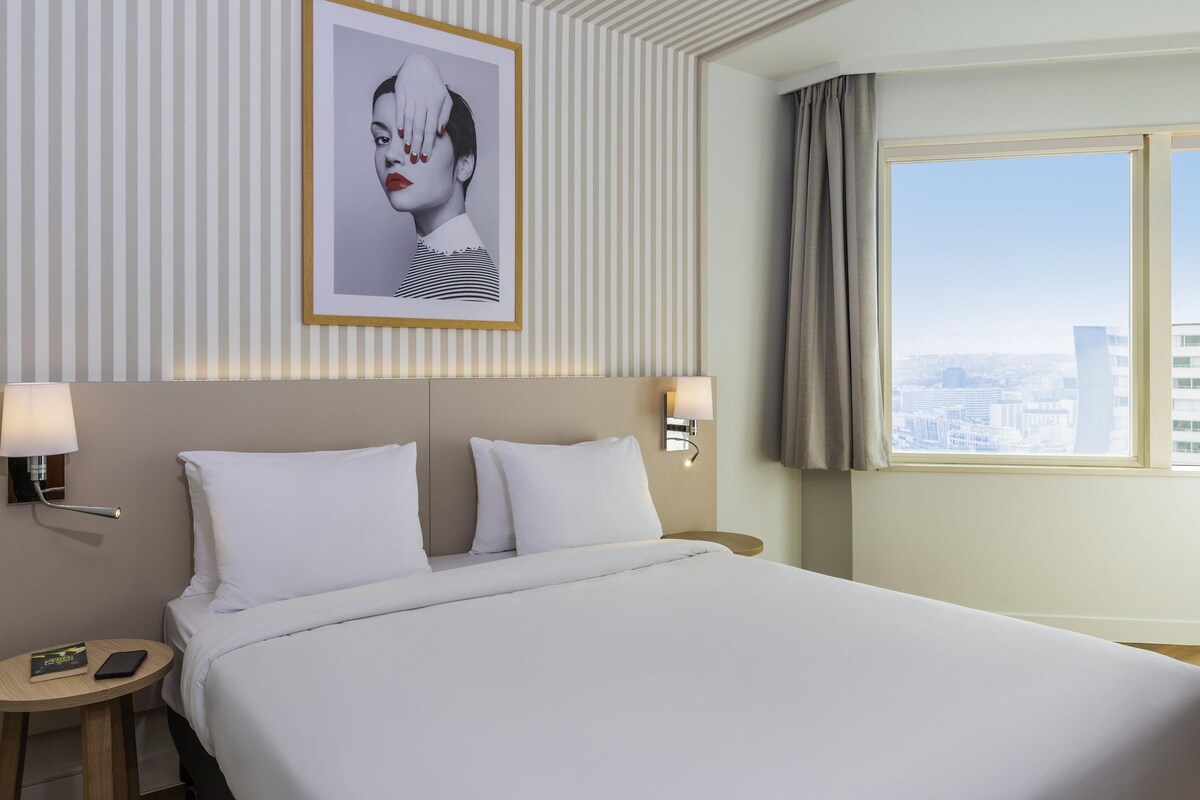 Una camera d'albergo con un letto, due comodini, un tavolino di fianco al letto e una finestra con vista sulla città.