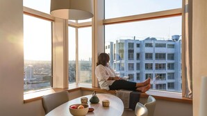 Una persona sentada junto a un amplio ventanal con vista a la ciudad, una mesa con un bol y una tetera, y una lámpara.