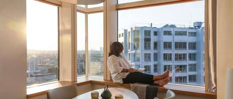 A person sitting by a large window with a city view, a table with a bowl and teapot, and a lamp.