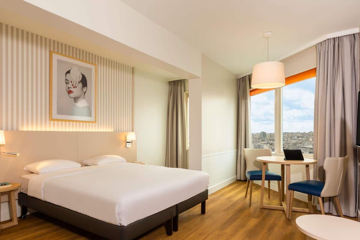 Habitación de hotel con una cama grande, un escritorio con una silla, una ventana con vista a la ciudad y una obra de arte colgada en la pared.