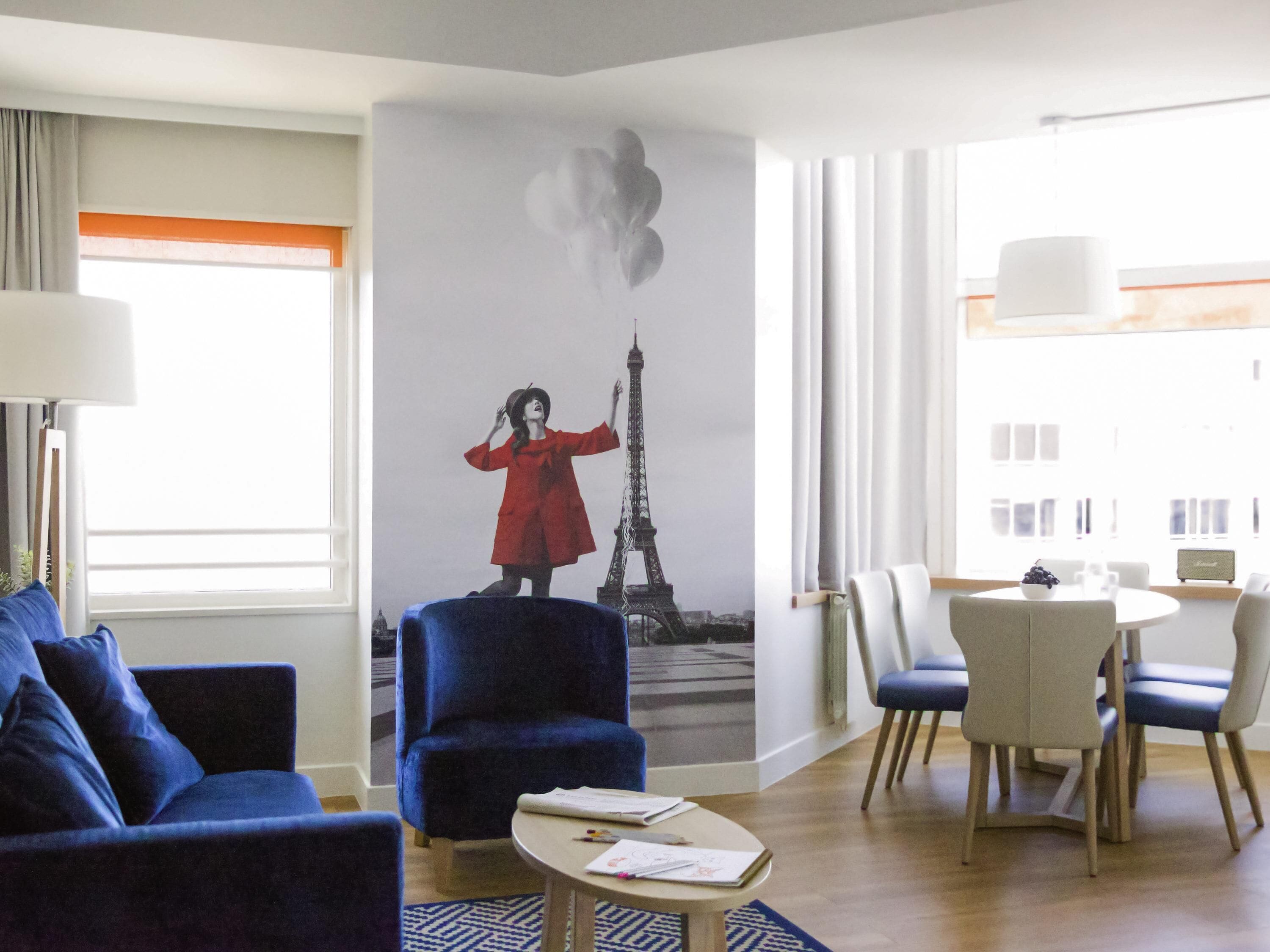 A hotel room with a large mural of the Eiffel Tower, a blue armchair, a round table with a white bowl, and a dining area with chairs.