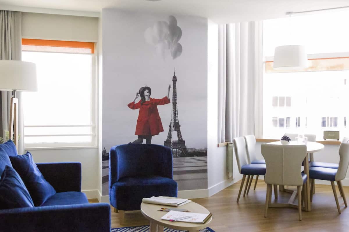 A hotel room with a large mural of the Eiffel Tower, a blue armchair, a round table with a white bowl, and a dining area with chairs.