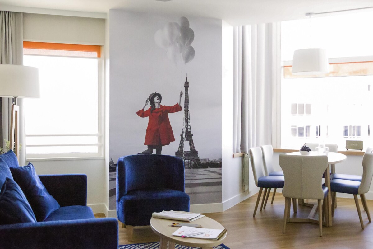 Habitación de hotel con un gran mural de la Torre Eiffel, un sillón azul, una mesa redonda con un bol blanco y una zona de comedor con sillas.