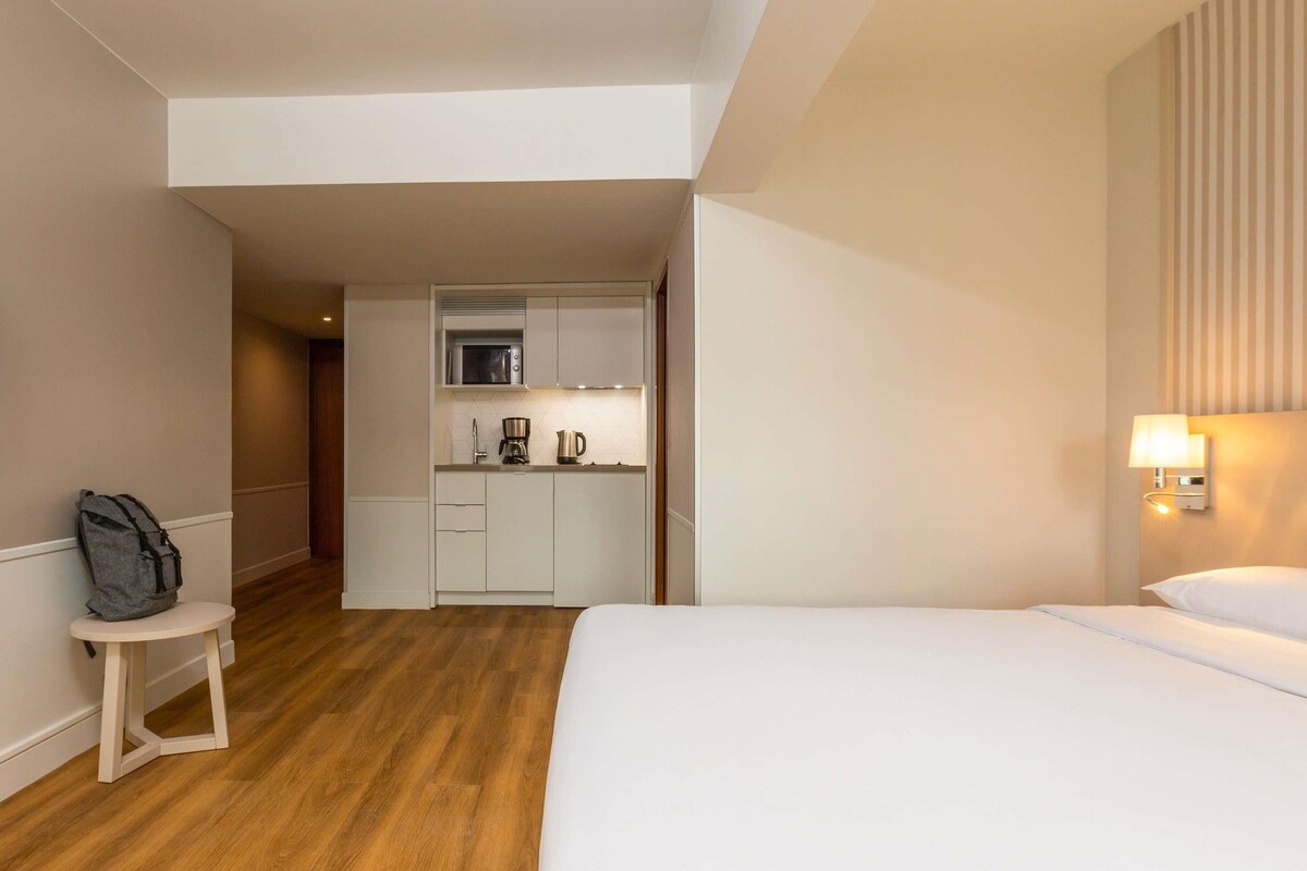 Camera d'albergo con un letto, un tavolino con uno zaino, angolo cottura con microonde e una lampada a parete.