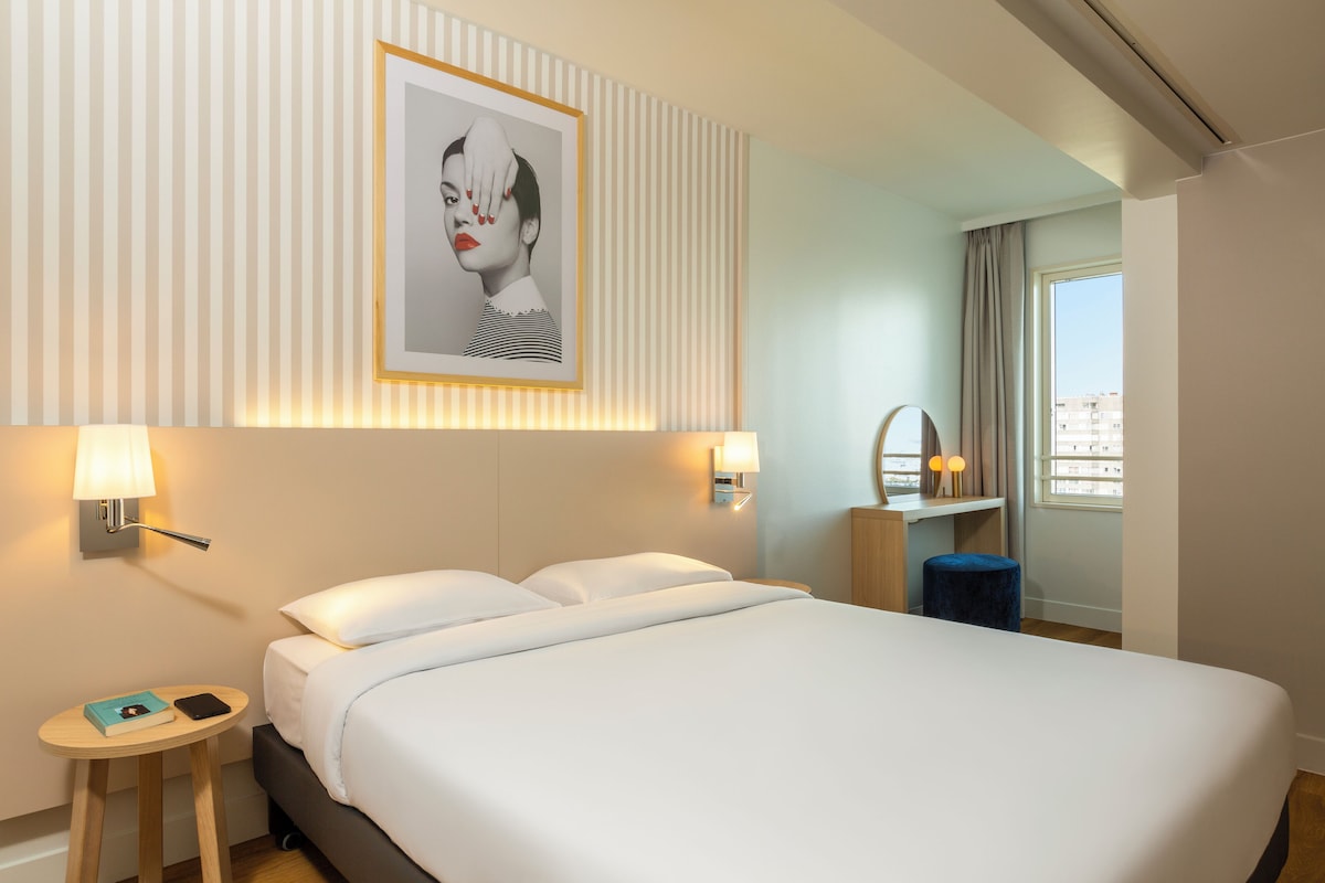 Una cama bien tendida con sábanas blancas, una mesita de noche con lámpara, un cuadro en la pared y una ventana con vistas a edificios.
