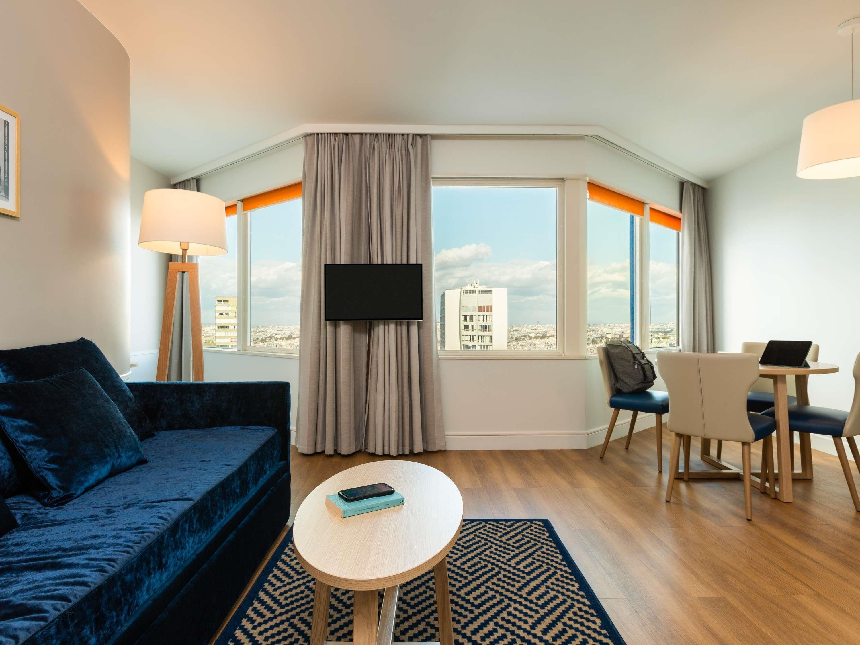 Quarto de hotel com sofá, mesa pequena, televisão e vista para a cidade.