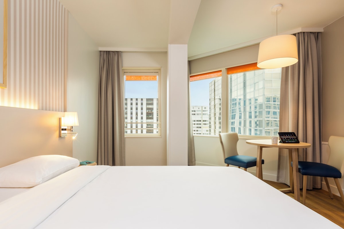 Habitación de hotel con cama, escritorio con silla, lámpara y vista de edificios a través del ventanal.