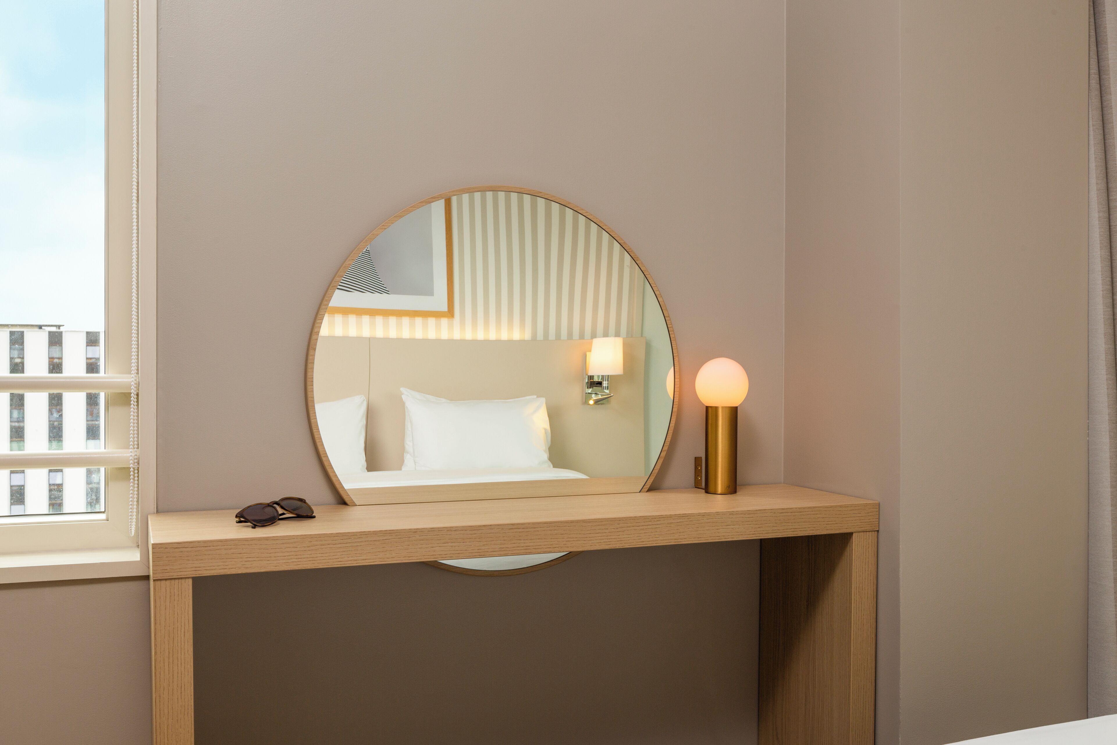 Une chambre d’hôtel avec un miroir rond, une lampe de chevet, un lit avec une literie blanche et une fenêtre avec des stores.