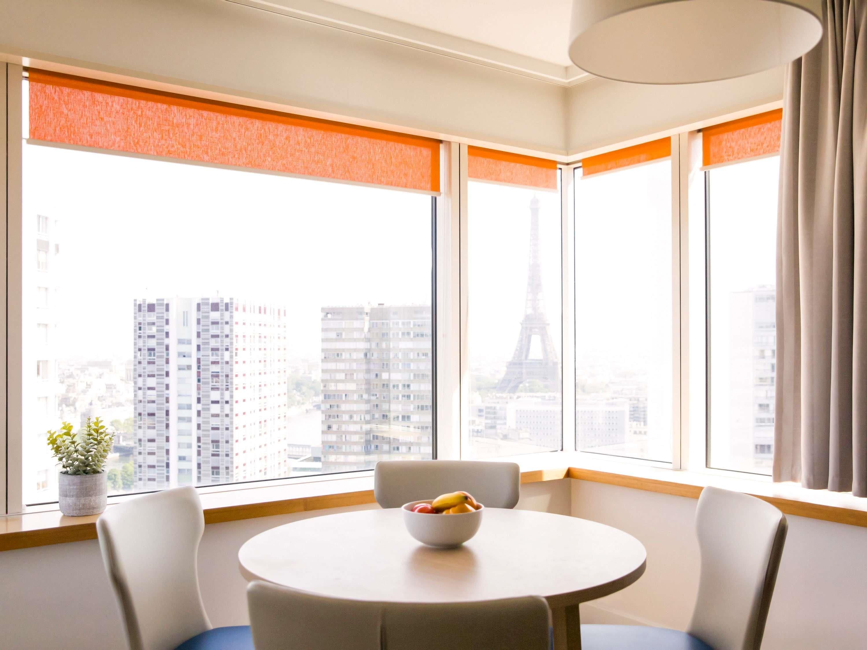 A modern hotel room with a round table and chairs, large windows with orange blinds, and a view of the Eiffel Tower.