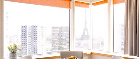 A modern hotel room with a round table and chairs, large windows with orange blinds, and a view of the Eiffel Tower.