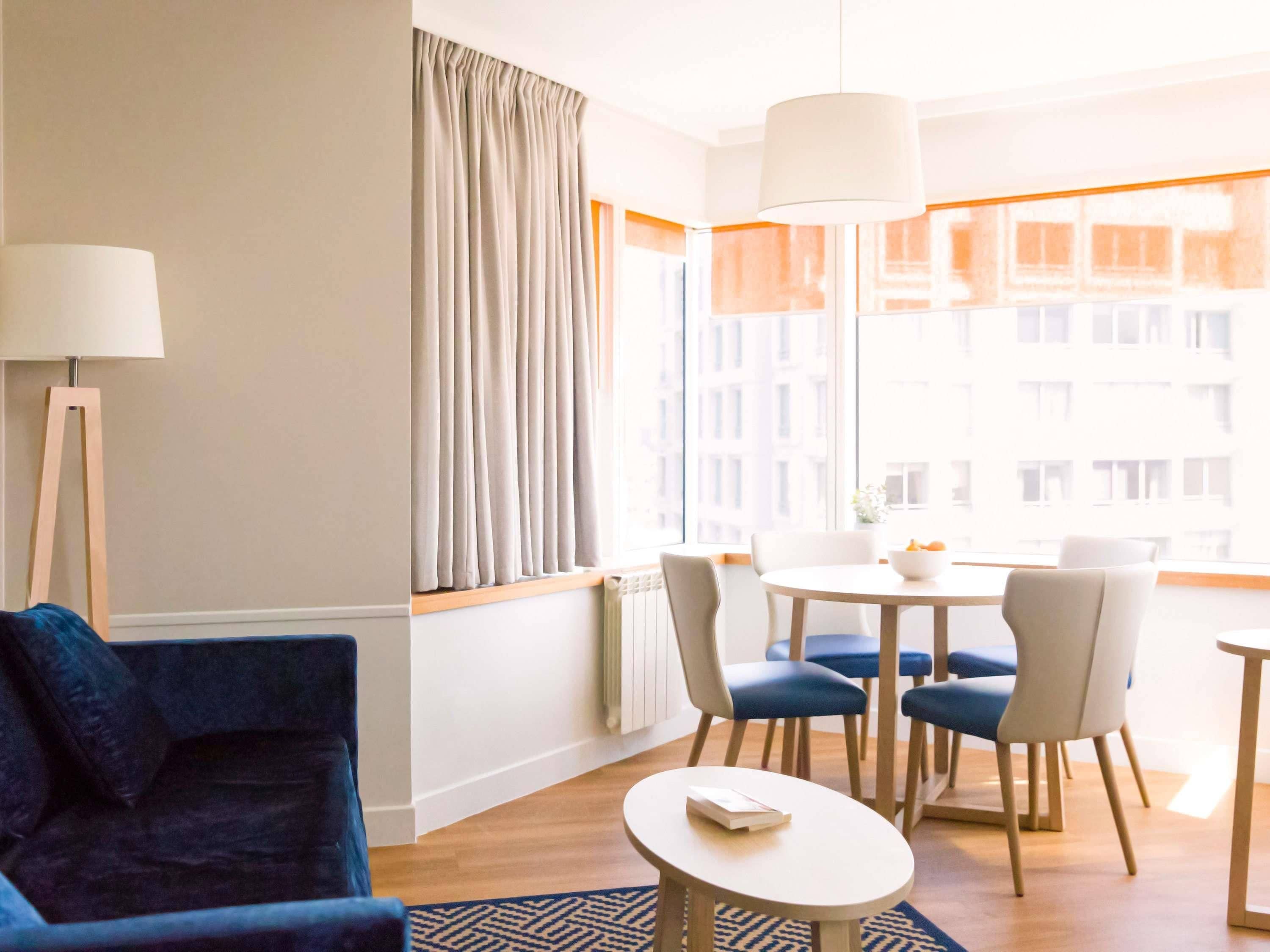 A hotel room with a blue armchair, a round table with chairs, a large window with curtains, and a hanging light fixture.