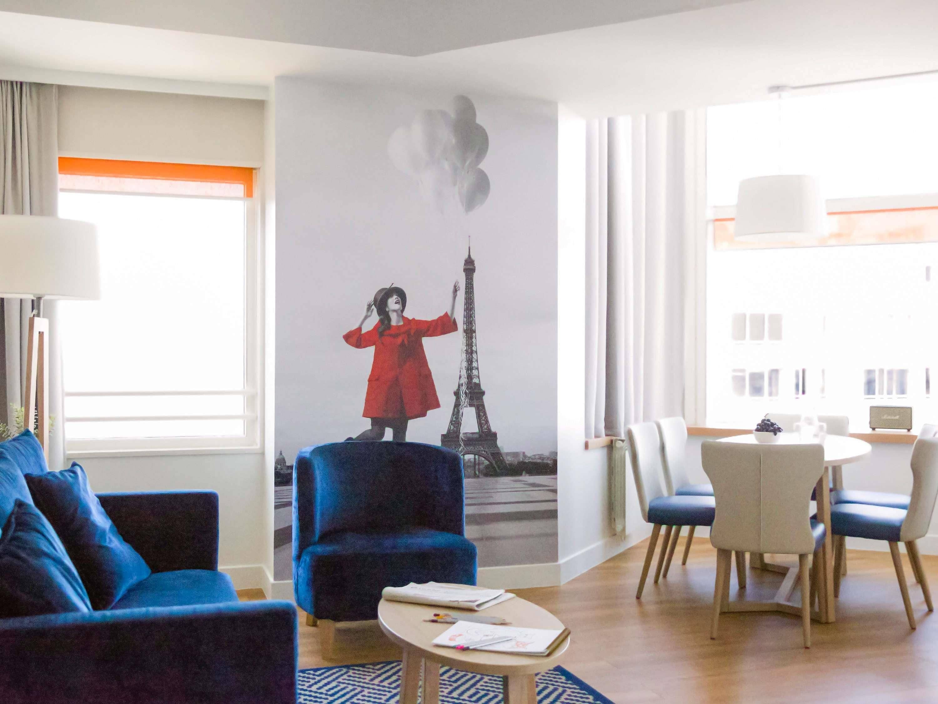 A hotel room with a large mural of the Eiffel Tower, a blue sofa, a dining table with chairs, and a small coffee table.