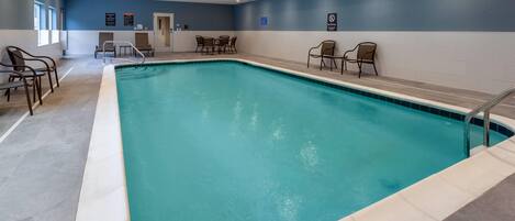 Una piscina interior con borde de azulejos, rodeada de sillones de descanso y una zona de estar con mesas.