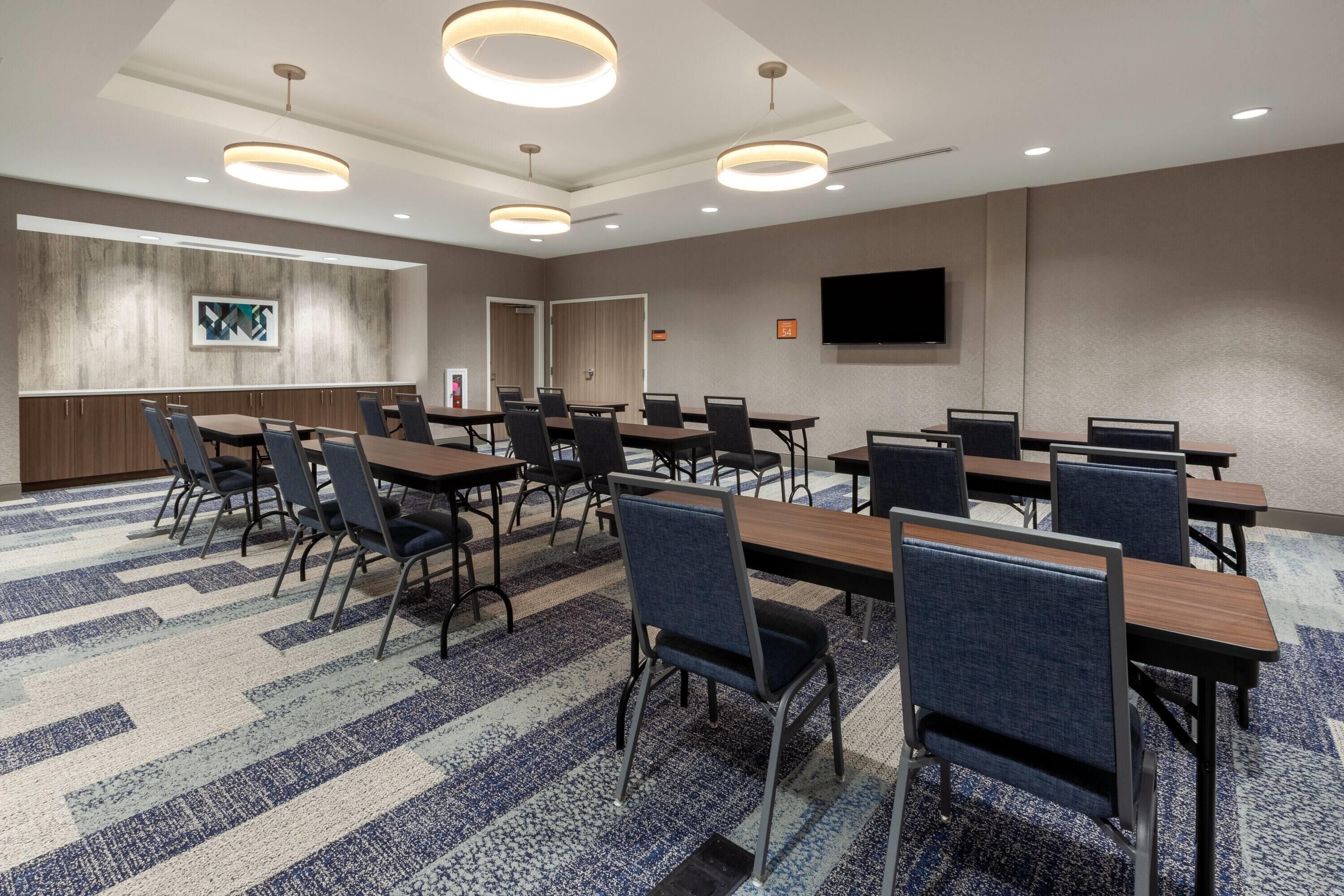 A conference room with multiple tables and chairs, a flat-screen TV mounted on the wall, and a carpeted floor.