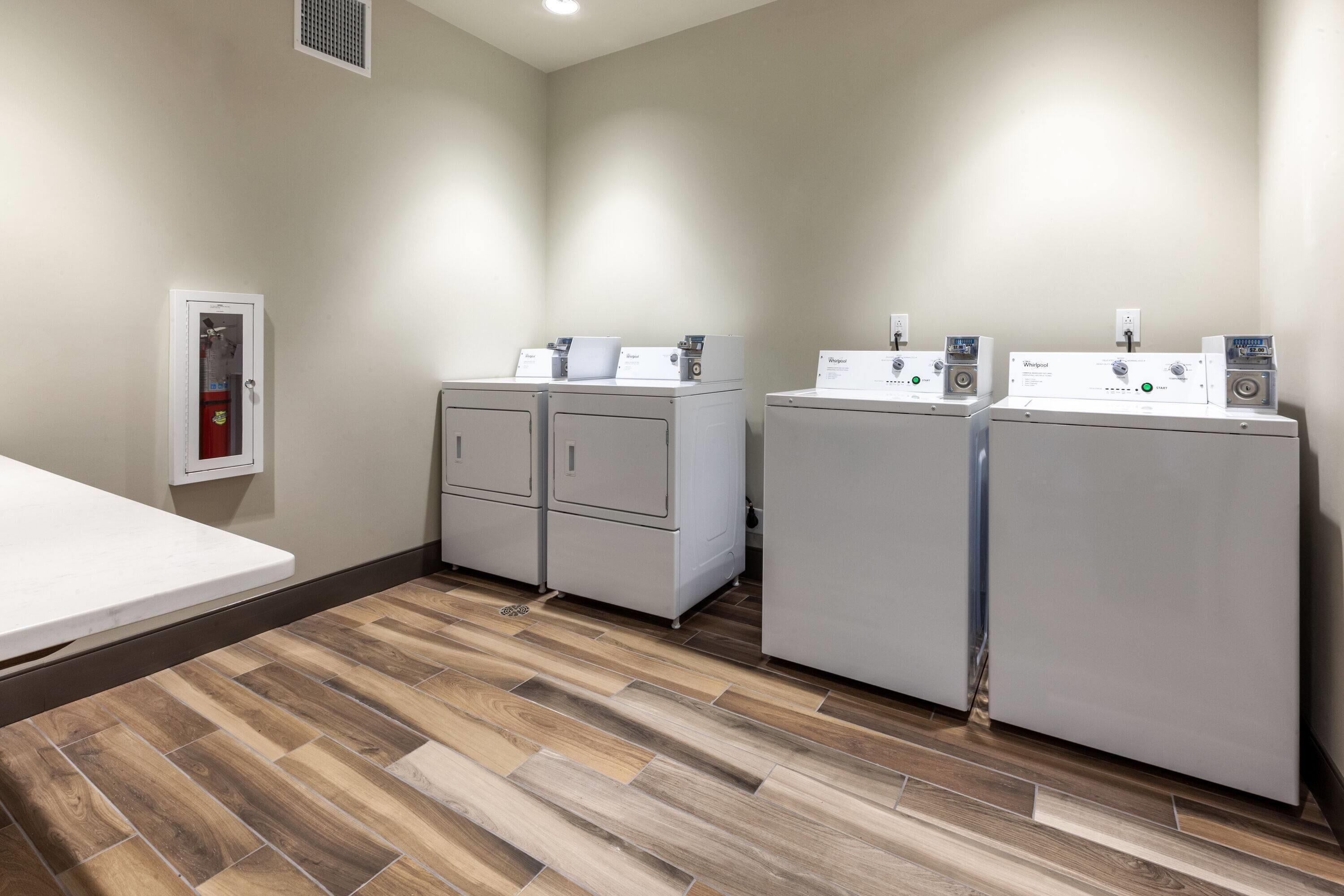 A laundry room with two Whirlpool washers and a dryer, a white table, and a fire extinguisher on the wall.