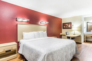 Una habitación de hotel con cama, mesitas de noche, un escritorio con silla, una lámpara y un cuadro en la pared.
