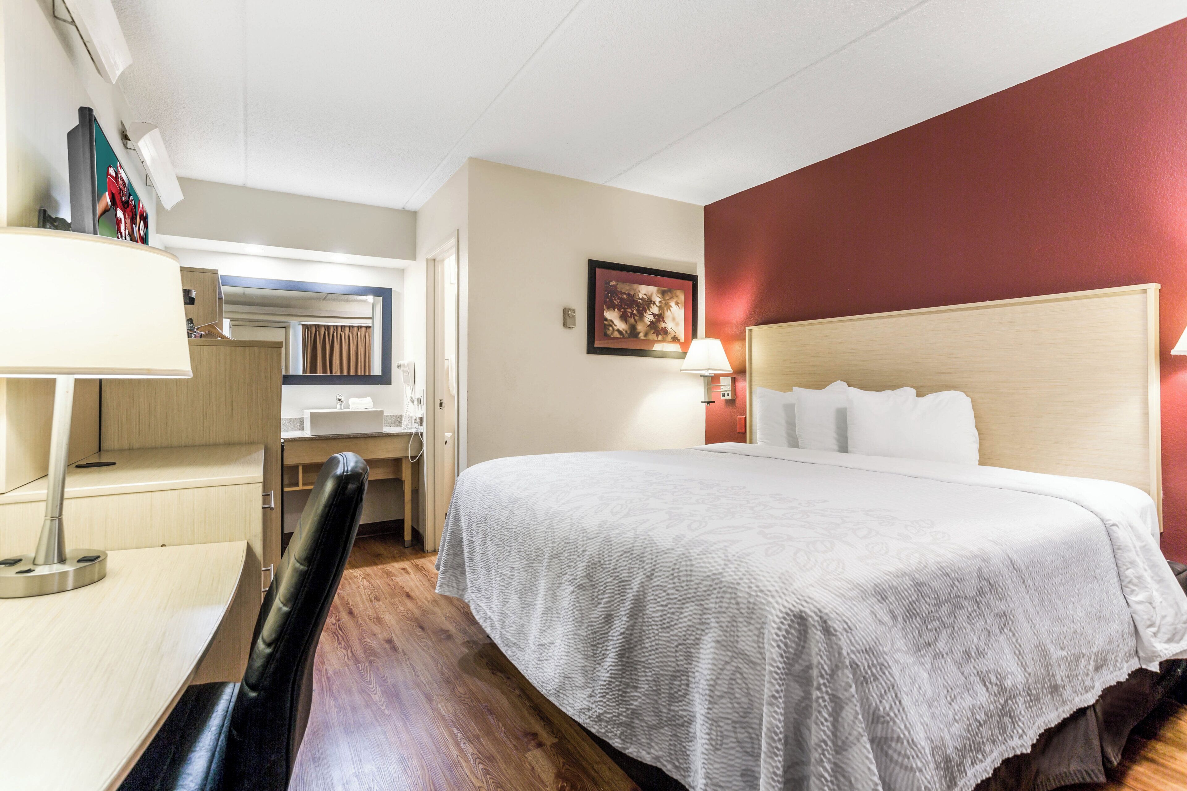 A hotel room with a bed, a desk, a chair, a lamp, a painting, and a window.