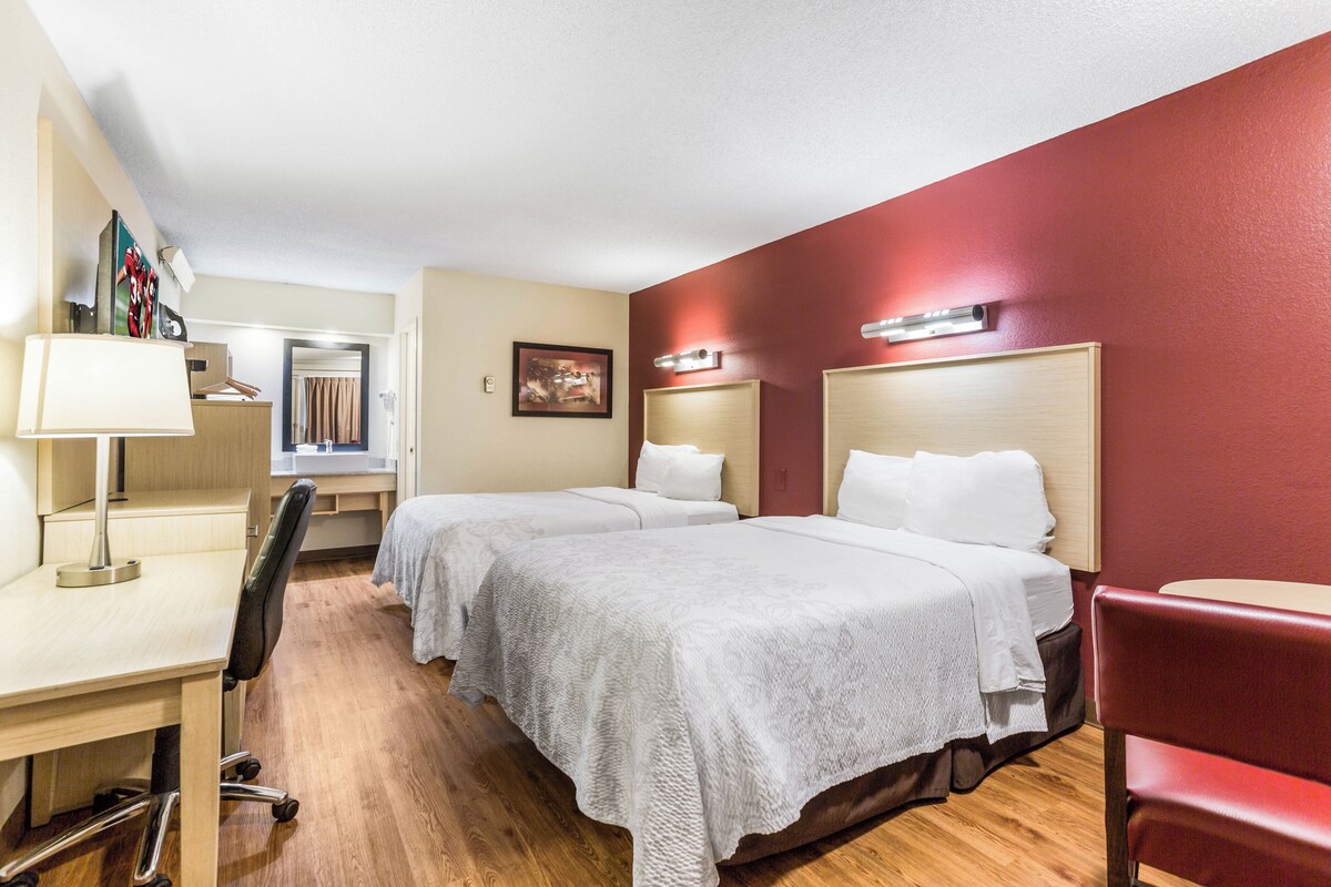 A hotel room with two beds, a desk, a chair, and a red accent wall.