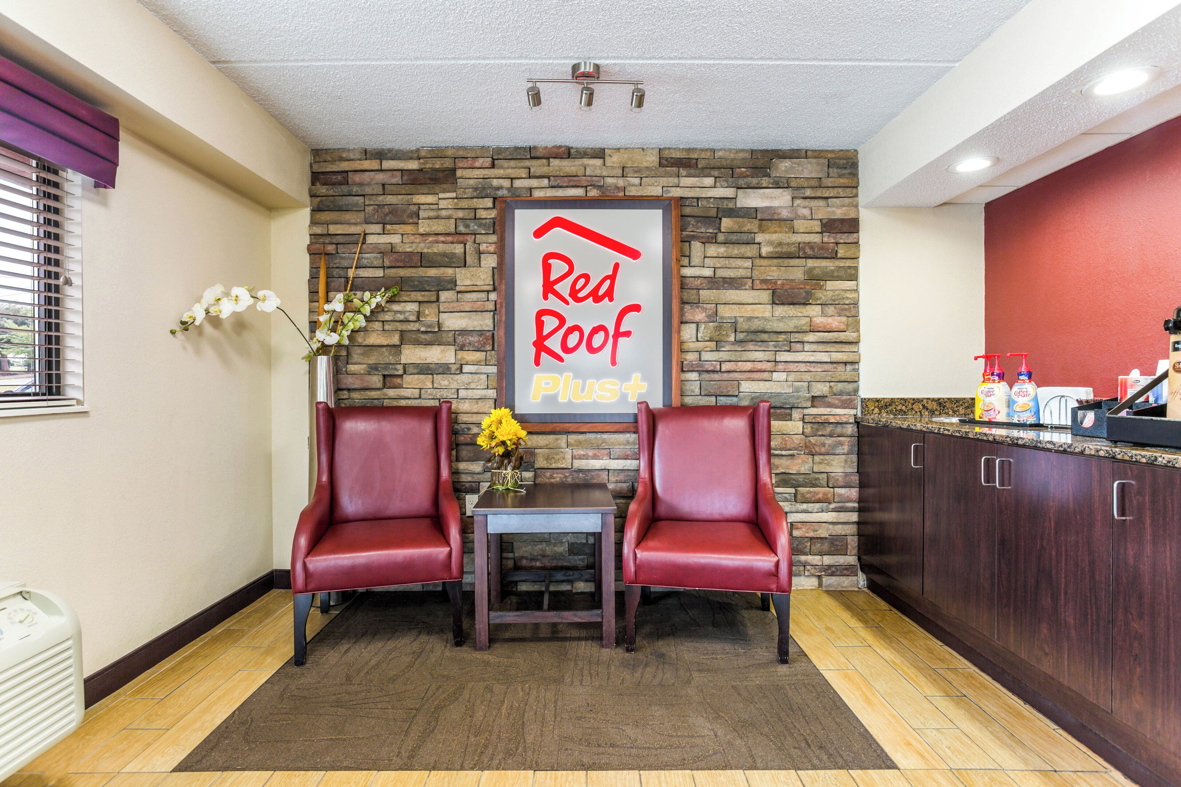Un hall d’accueil de l’hôtel Red Roof Plus, avec deux fauteuils en cuir, une petite table, un vase de fleurs et un mur en pierre en arrière-plan.