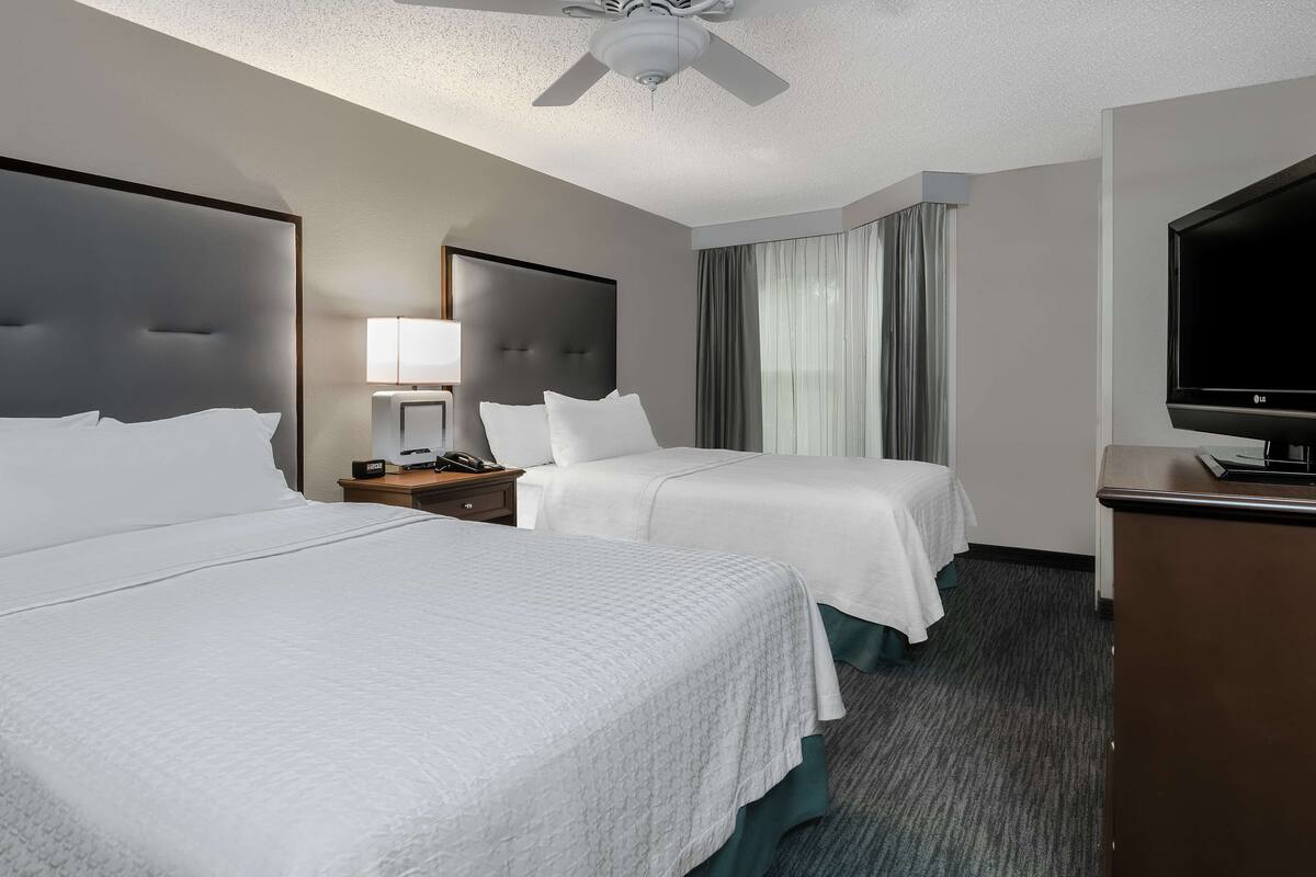 A hotel room with two beds, a ceiling fan, a TV, and a desk with a lamp.