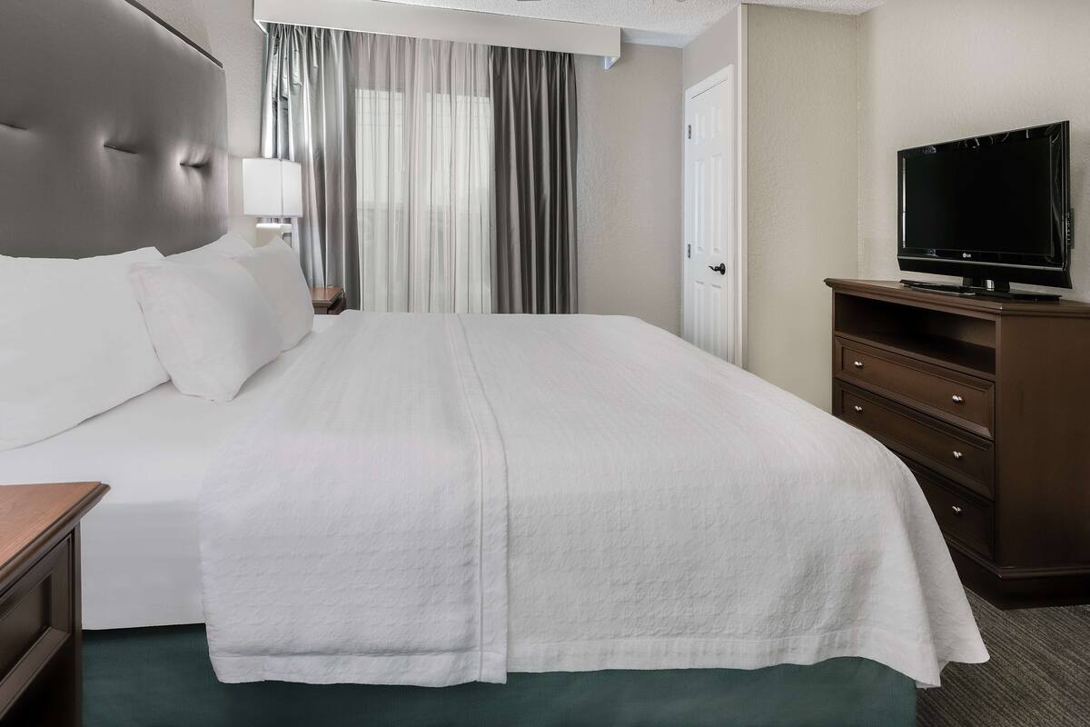 A neatly made bed with white linens, a television on a wooden stand, and a window with curtains.