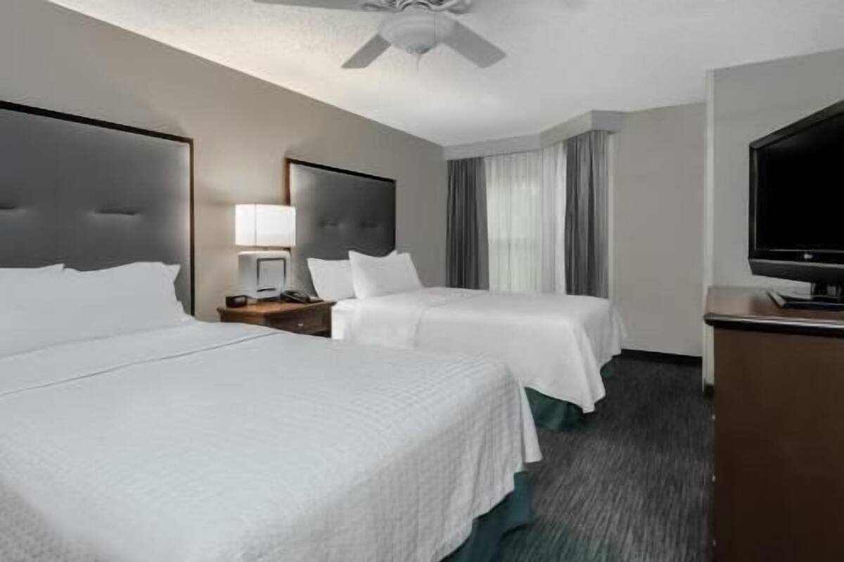 A hotel room with two beds, a desk with a lamp, a television, and a ceiling fan.