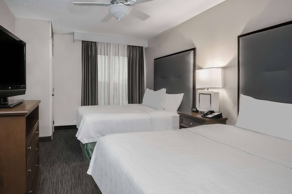 A hotel room with two beds, a TV, a ceiling fan, a desk with a lamp, and a large mirror.