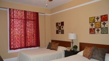 A hotel room with two beds, a window with red curtains, a bedside table with a lamp, and wall art.