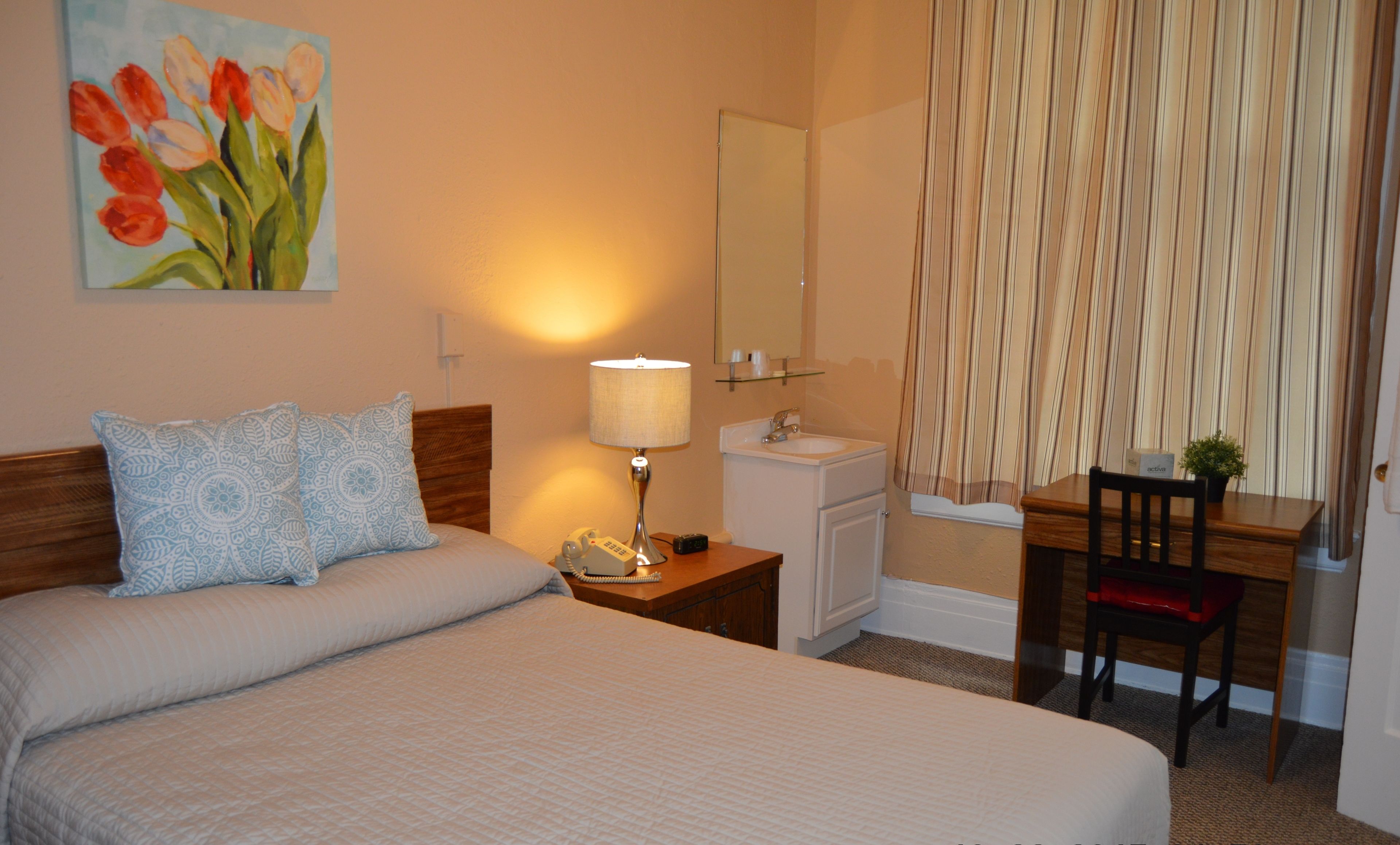 A hotel room with a bed, a bedside table with a lamp, a desk with a chair, a bathroom, and a painting on the wall.