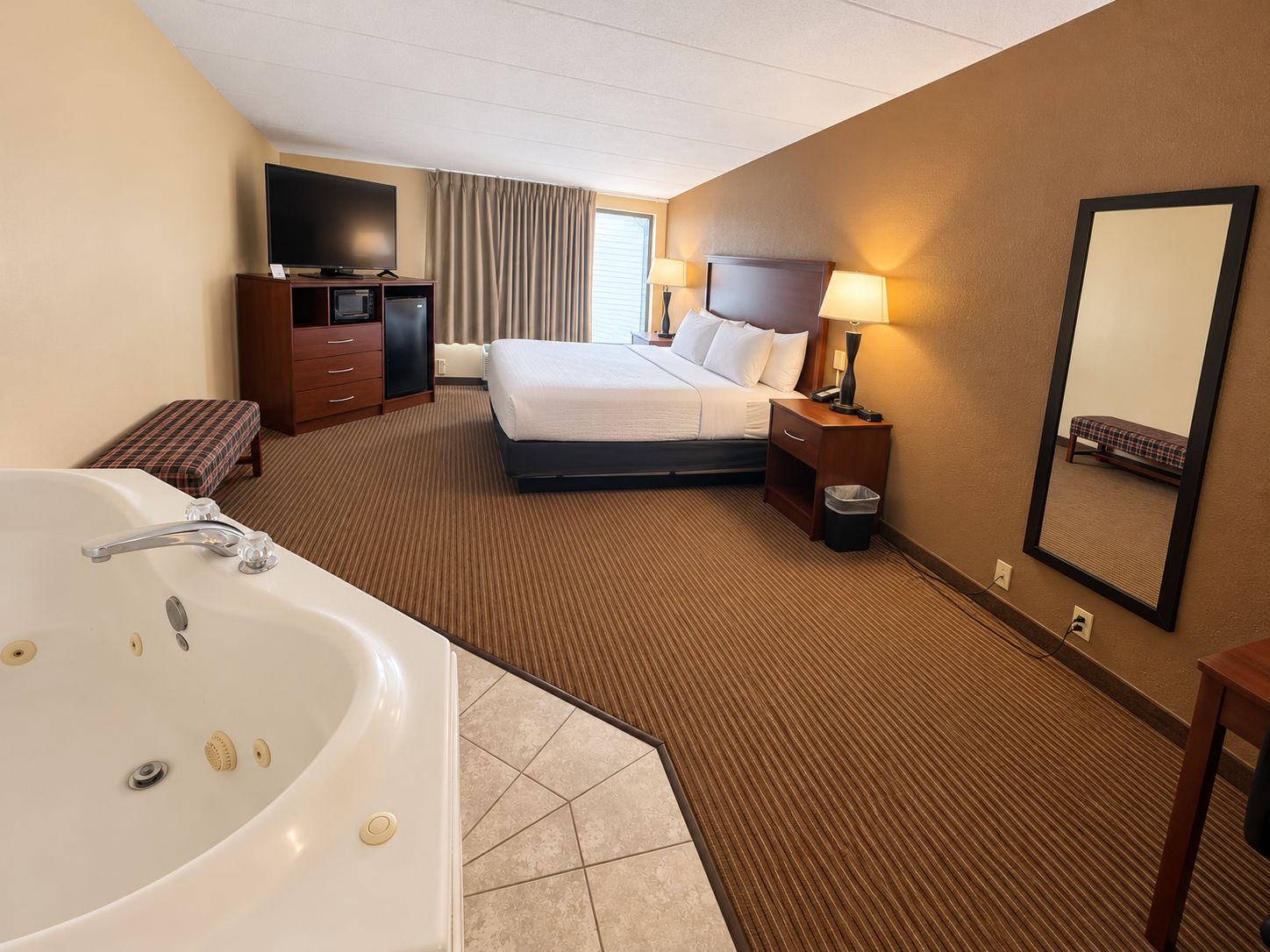 A hotel room with a large bed, a TV, a desk with a computer, a desk lamp, a bedside table with a lamp, a mirror, and a bathroom with a bathtub and sink.