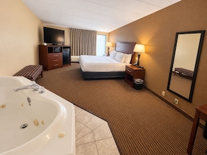 A hotel room with a large bed, a TV, a desk with a computer, a desk lamp, a bedside table with a lamp, a mirror, and a bathroom with a bathtub and sink.