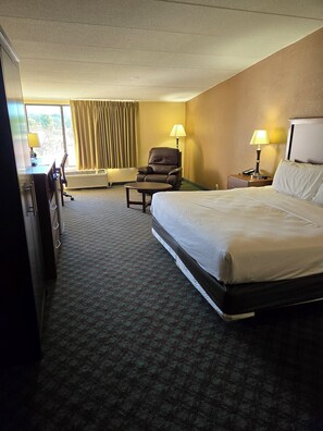 A hotel room with a large bed, a desk with a chair, a chair, two lamps, and a window with curtains.