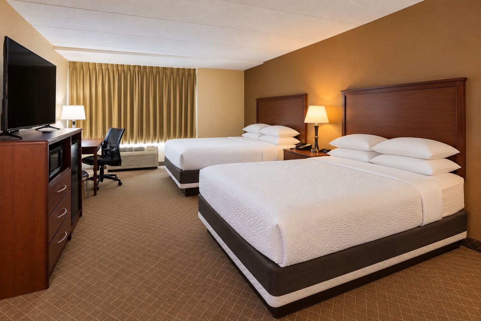 A hotel room with two beds, a desk, a chair, a television, and a lamp.
