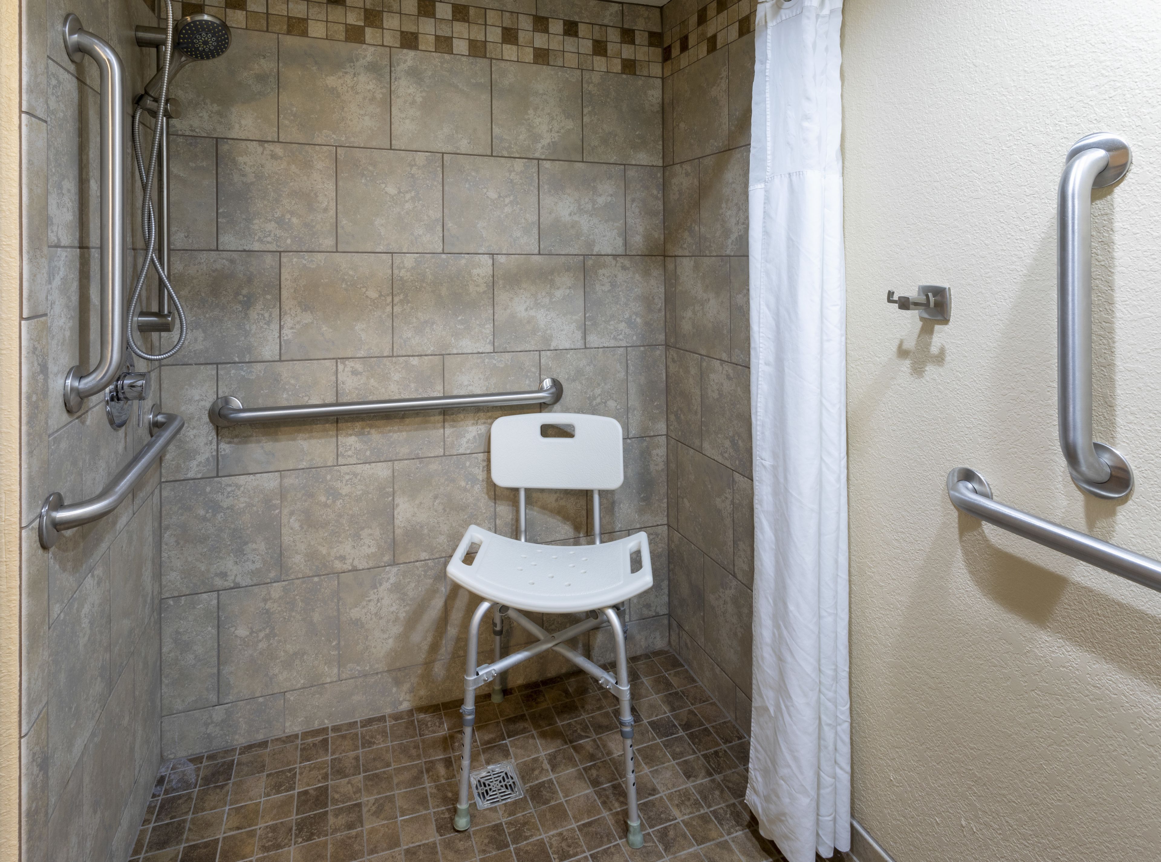 Un espace douche équipé de barres d’appui, d’une chaise blanche et d’un pommeau de douche.