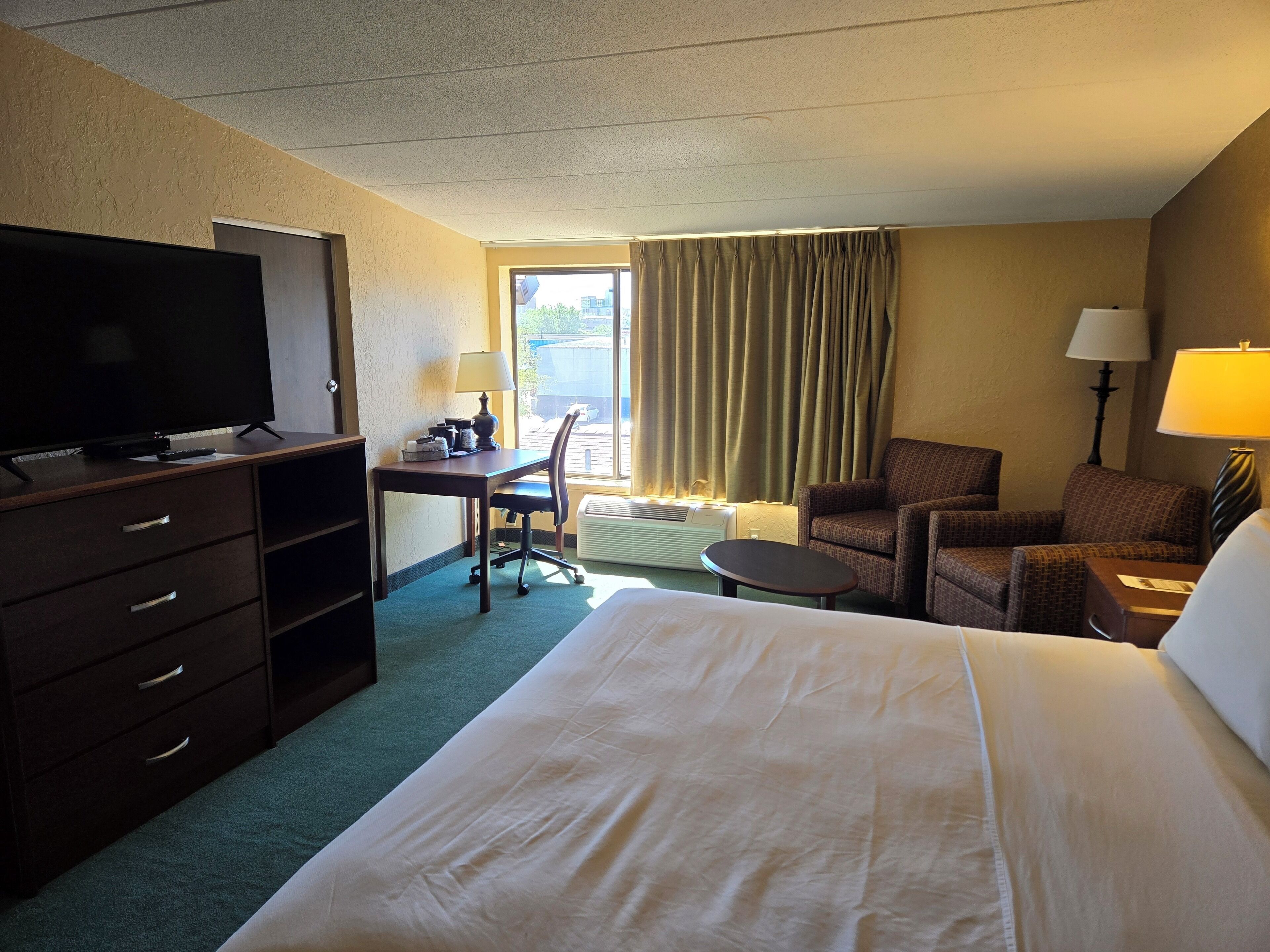 A hotel room with a bed, a desk with a chair, a television, a lamp, and a window with curtains.