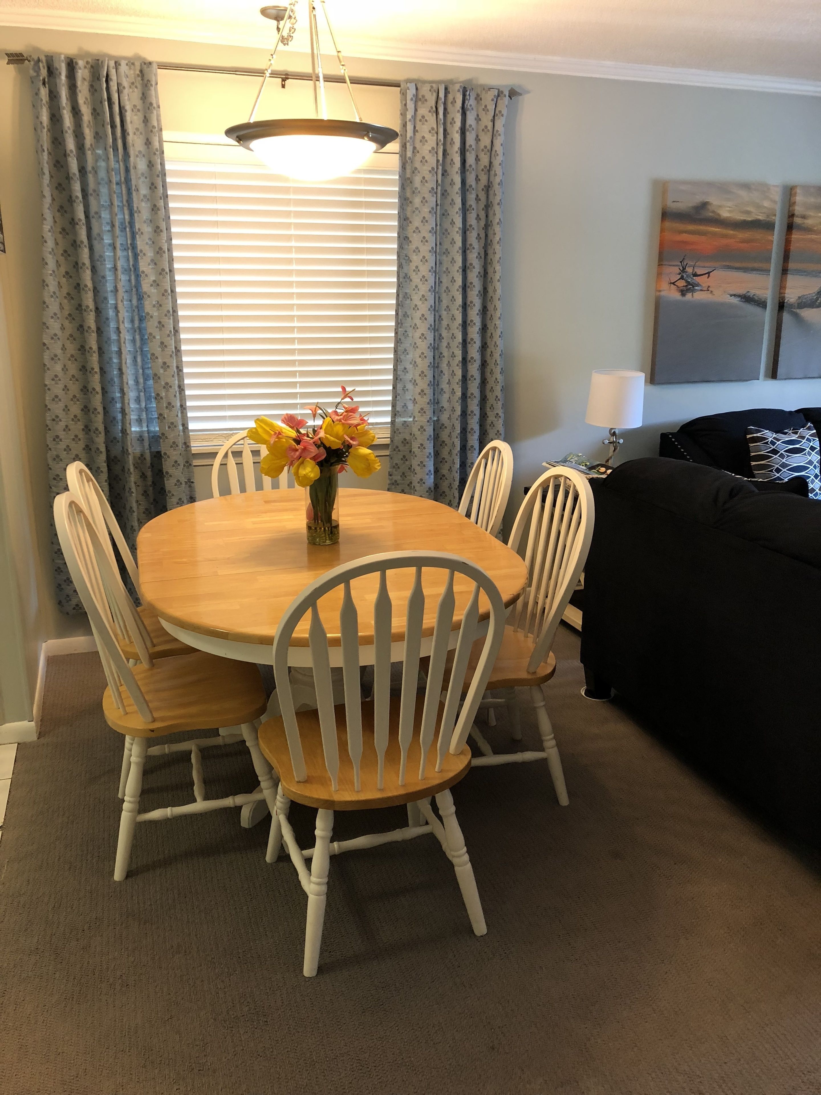 Een eetruimte met een ronde houten tafel en witte stoelen, een vaas met bloemen, en een schilderij aan de muur.