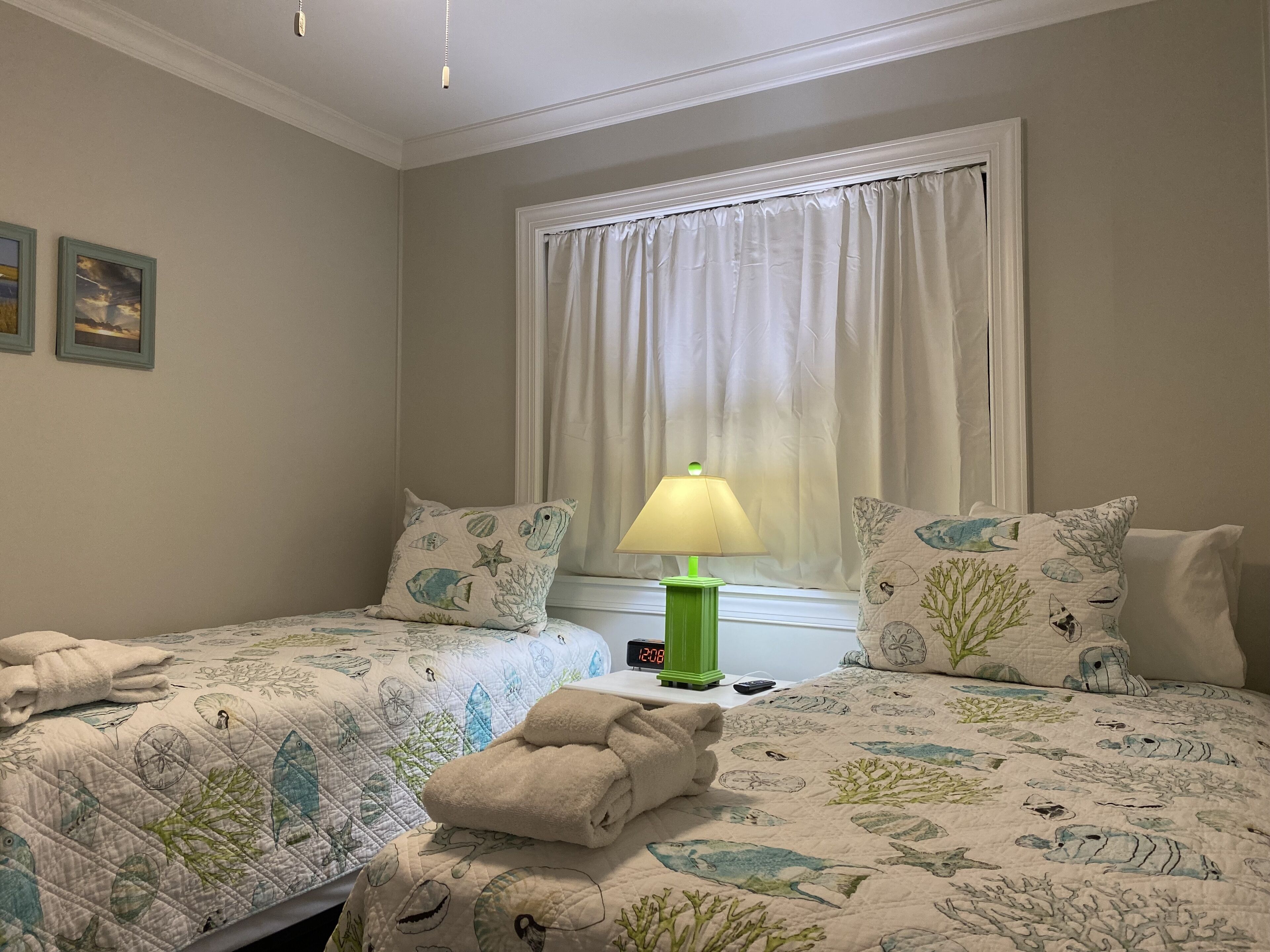 Un dormitorio con dos camas, una mesita de noche, una lámpara verde y una ventana con cortinas.