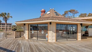 Une terrasse en bois surmontée d’une structure fermée par des parois de verre, probablement un salon ou un espace d’attente, offrant une vue dégagée sur les bâtiments environnants et les palmiers.