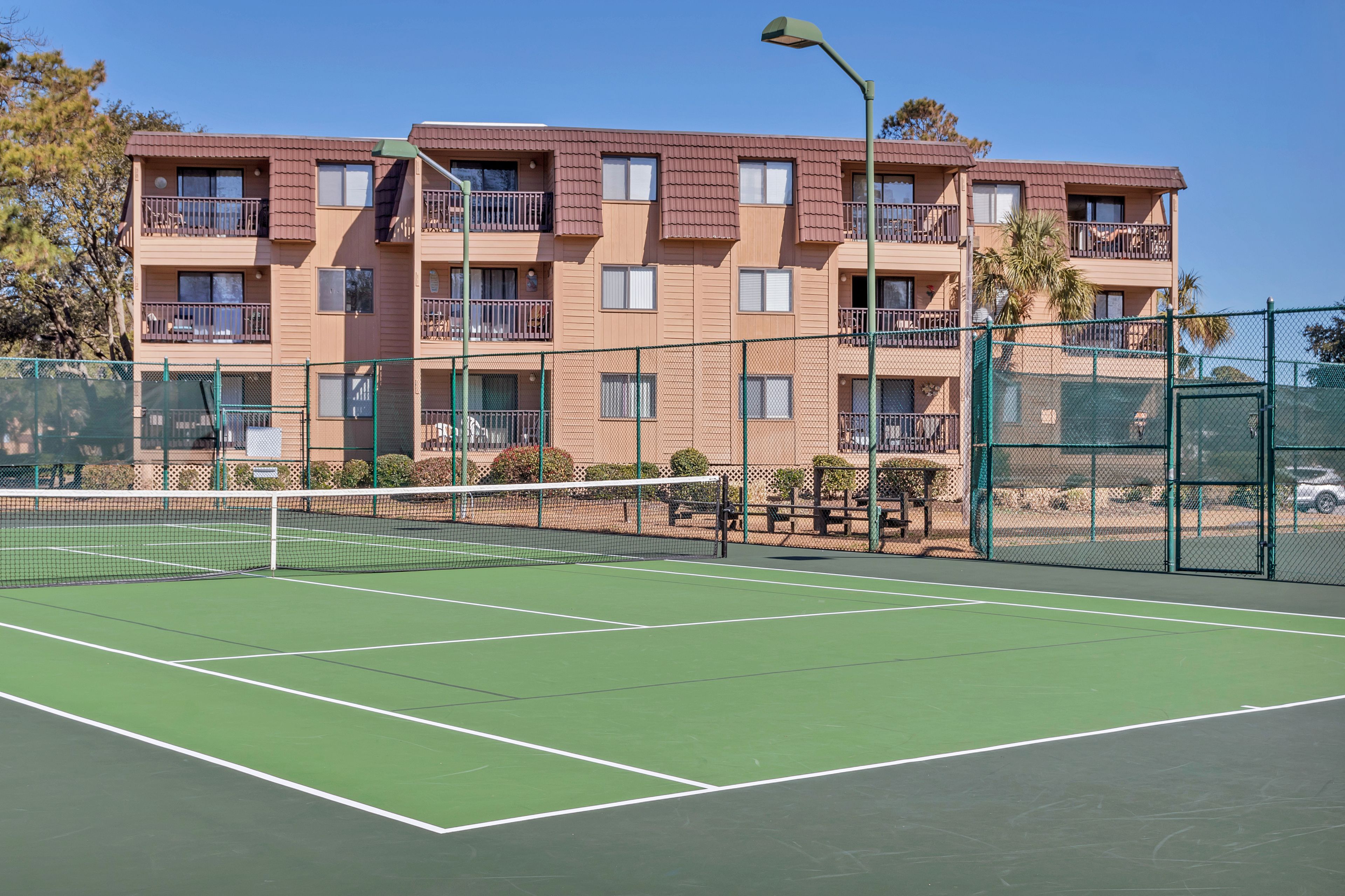 A tennis court adjacent to a multi-story building with balconies and a green fence.