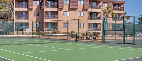 A tennis court adjacent to a multi-story building with balconies and a green fence.