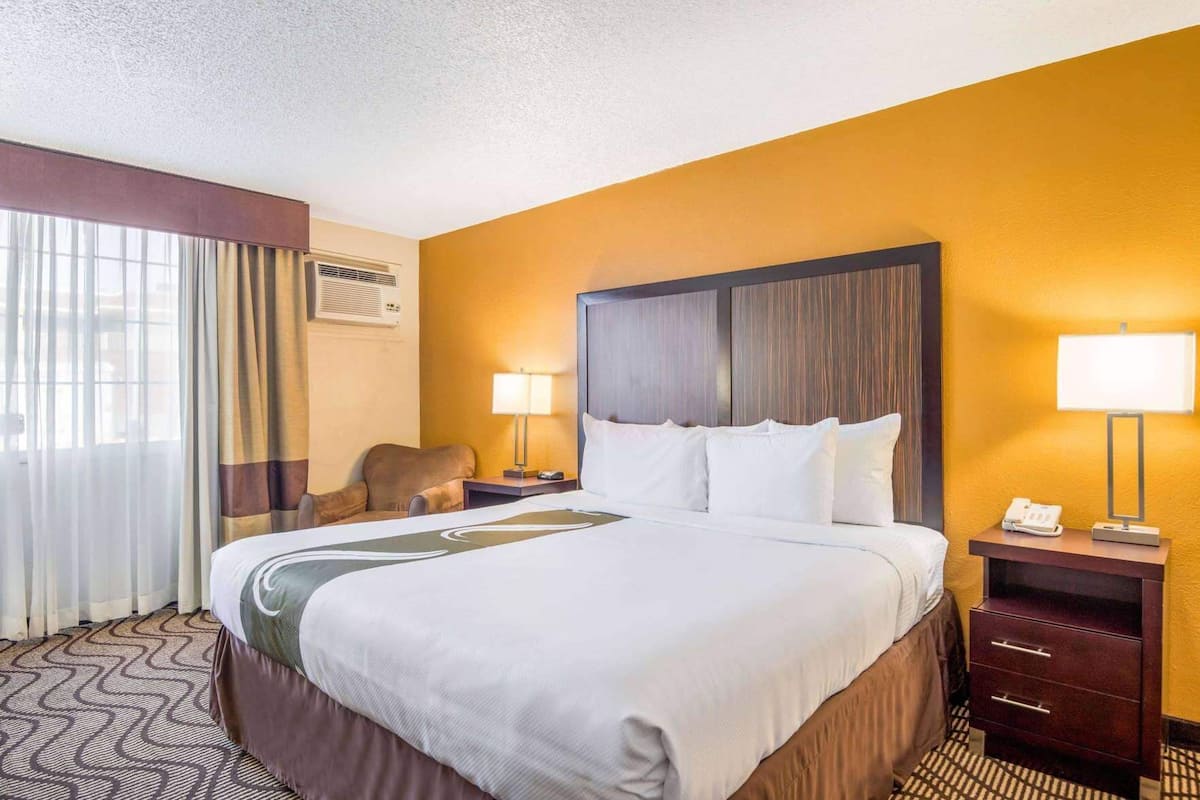 A hotel room with a large bed, a desk with a lamp, a chair, and an air conditioning unit.