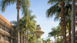 Una fuente de varios niveles con esculturas decorativas, rodeada de palmeras y una piscina, ubicada en un patio con un edificio al fondo.
