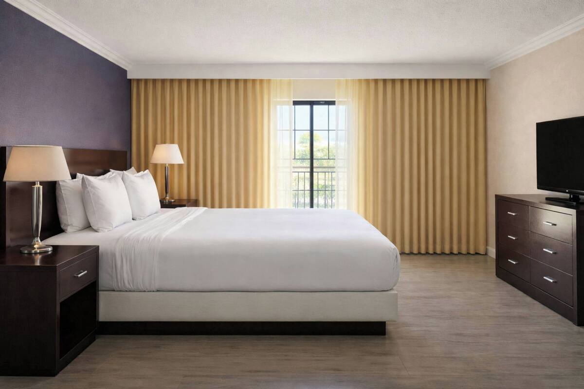 Una habitación de hotel con cama, televisión, mesita de noche, lámpara y ventana con cortinas.
