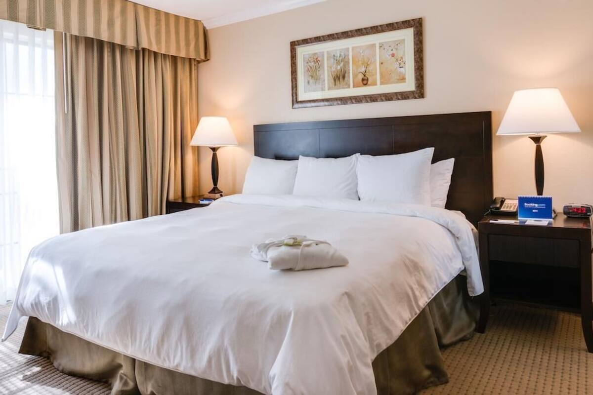 A hotel room with a large bed, two bedside lamps, a framed artwork above the bed, and a clock on the nightstand.