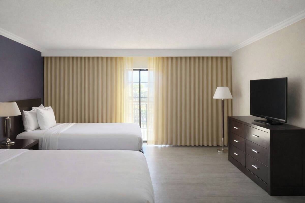 Habitación de hotel con dos camas, un televisor, una lámpara y un ventanal grande con cortinas.