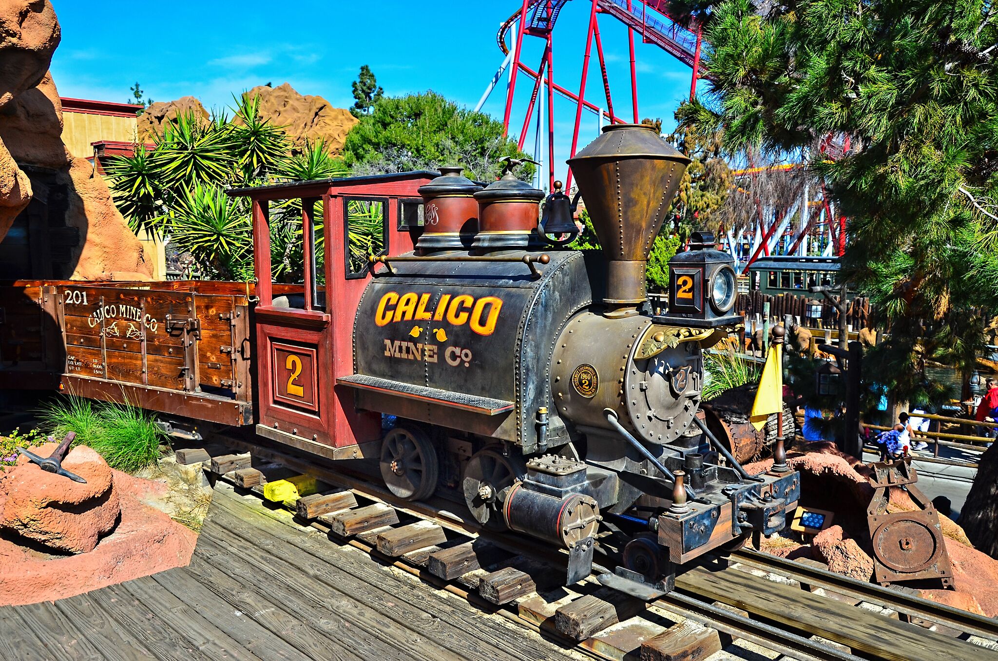 Un tren antiguo con la inscripción "CALICO MINE CO" y el número "2", circulando sobre raíles, con una plataforma de madera y terreno rocoso a su alrededor.