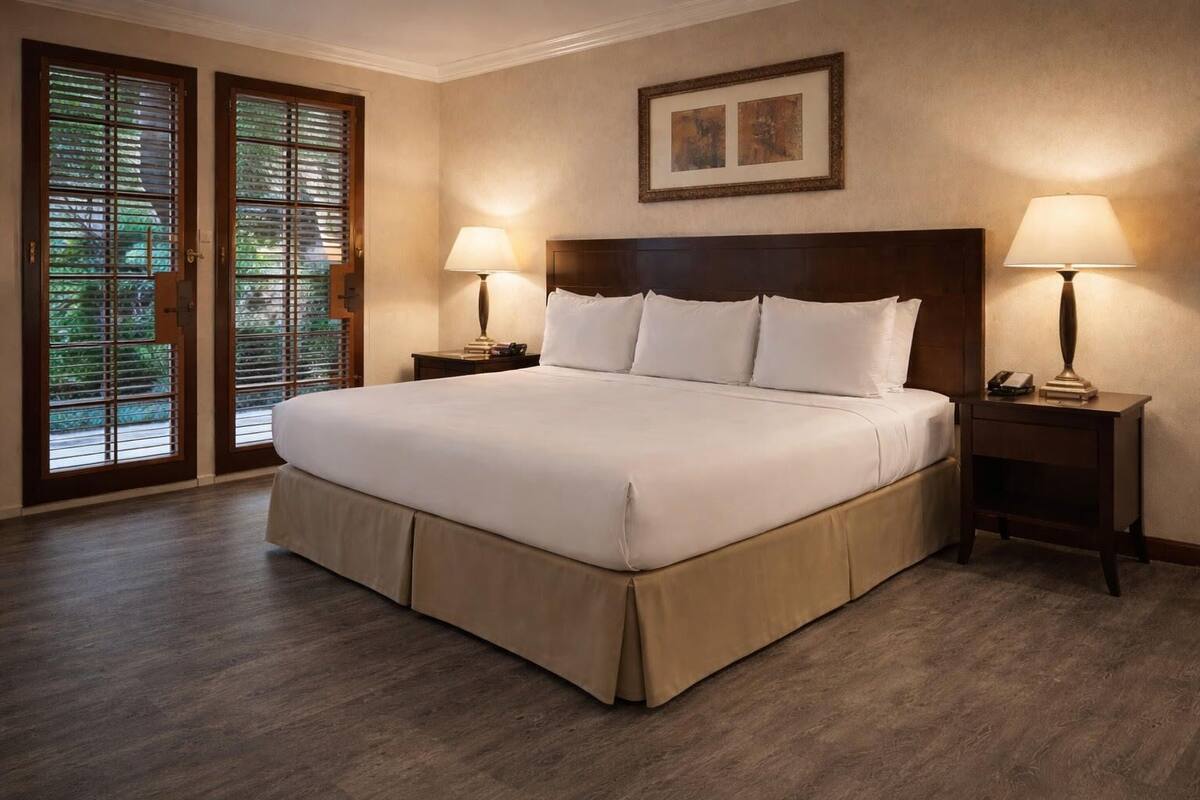 Una cama grande con cabecera de madera, dos mesitas de noche con lámparas, un cuadro enmarcado en la pared y una puerta de vidrio corredera con persianas.